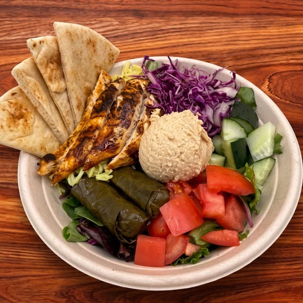 A Mediterranean bowl with grilled chicken, pita, hummus, dolmas, and fresh vegetables on a wooden table.